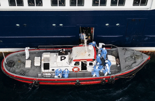 An aerial view shows health personnel assisting patients onto a boat from the cruise ship MV Hondius. Medics in blue suits can be seen guiding passengers off the ship onto a smaller boat for evacuation