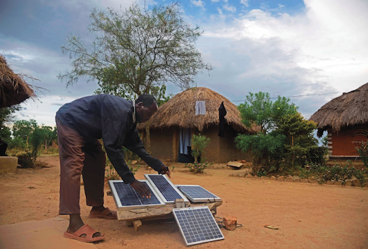 Person adjusting solar panels outside thatched-roof homes in a rural village.