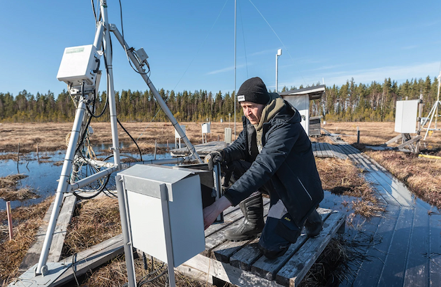 A researcher kneels before a metal box and contraption on top of a wooden pier in a grassy marsh.