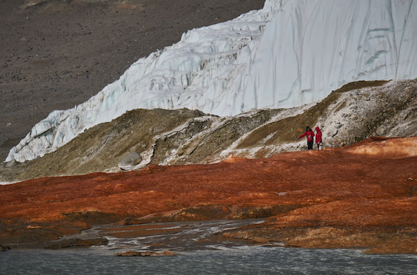 Two people in red coats stand in between rust-colored water and a glacier