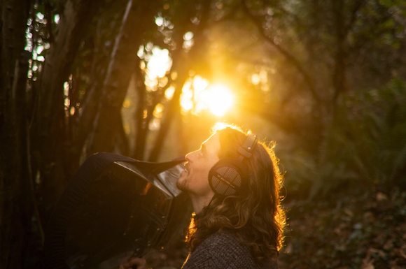A man wearing headphones stares up into the trees, backlit by the setting sun