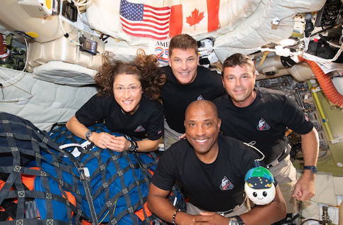 Four astronauts in black T shirts smiled for a photo inside their capsule.