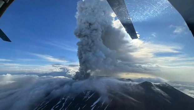 Airborne view of a column of ash erupting from a clouded mountain.