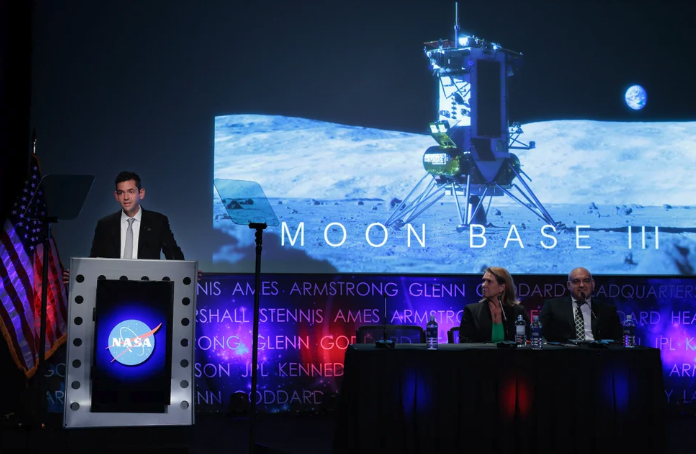Jared Isaacman stands behind a podium in front of a projector screen showing an image of a moon lander.