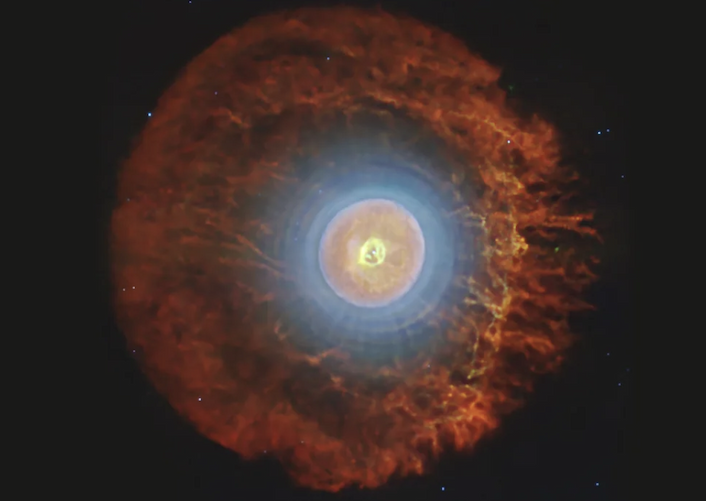 A ring-shaped nebula with a rust-colored out later and a bright yellow center containing a shape that resembles and upside down question mark.