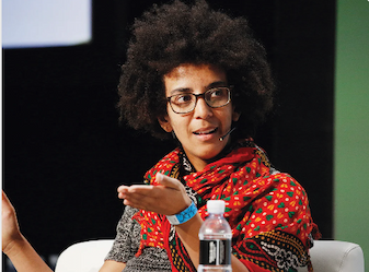 A photograph of Timnit Gebru wearing a bright read scarf,sitting in a chair gesticulating and wearing a microphone.