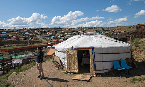 A man stands outside a white yurt-type structure on a bluff above a small village in an arid climate