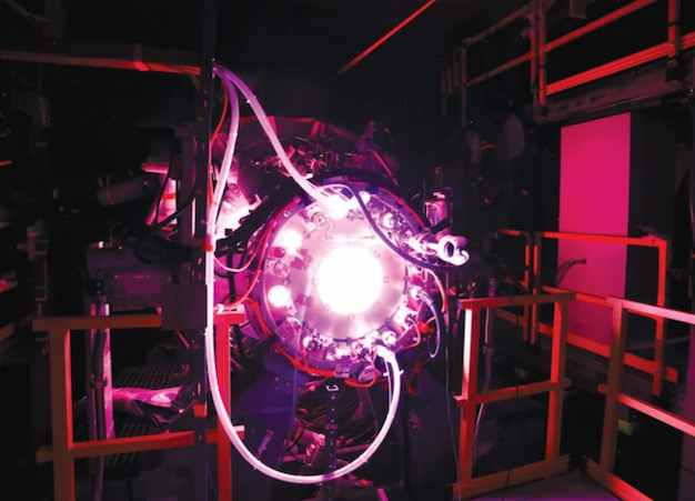 A cylindrical fusion prototype glows white and pink inside a dark test bay, surrounded by cables, hoses and metal supports.