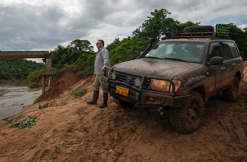 A man standing by a Jeep that drove on muddy dirt roads.
