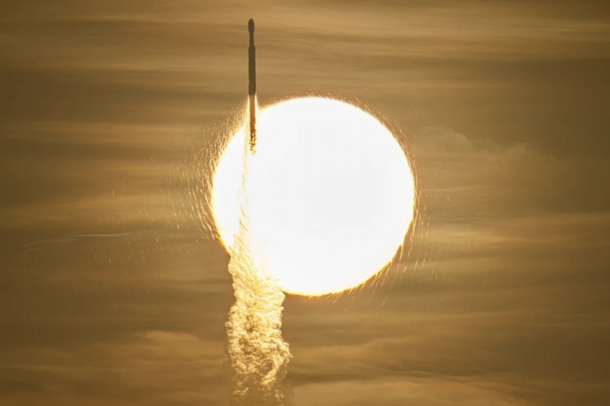 A rocket flies up in front of a bright sun, leaving smoke in its wake