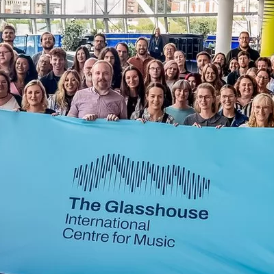 A crowd in The Glasshouse foyer all smile as they hold a blue flag with the new logo on for The Glasshouse International Centre for Music.