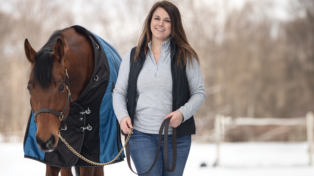 A rider leading her horse, wearing a blue turnout blanket, across a snowy field