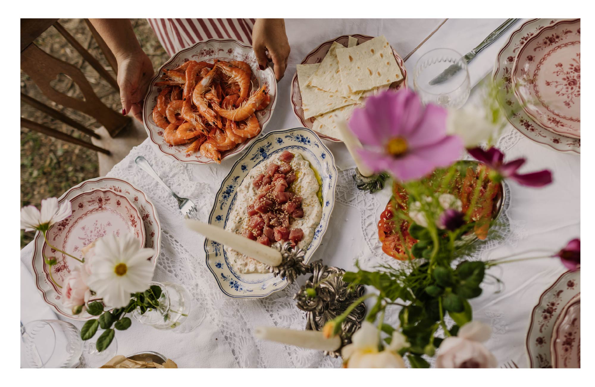 Photograph of an outdoor table with grilled shrimp, cheese, toast, a dip and Vila collection plates, decorated with flowers