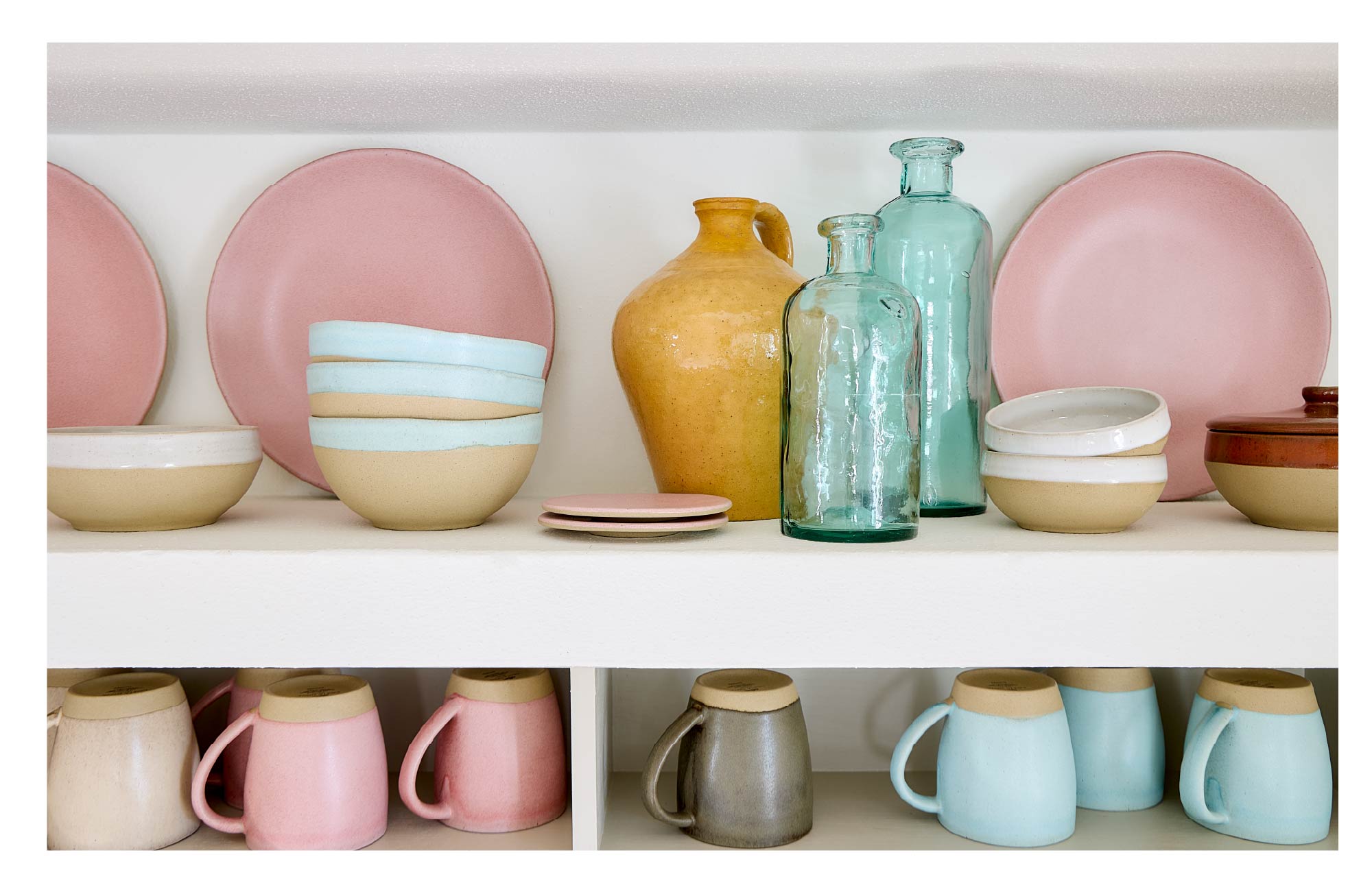 Shelves with pastel-toned dinnerware, pink plates, light blue and white bowls, amber and seafoam glass jugs and bottles, and pink, beige, light blue and grey mugs lined up