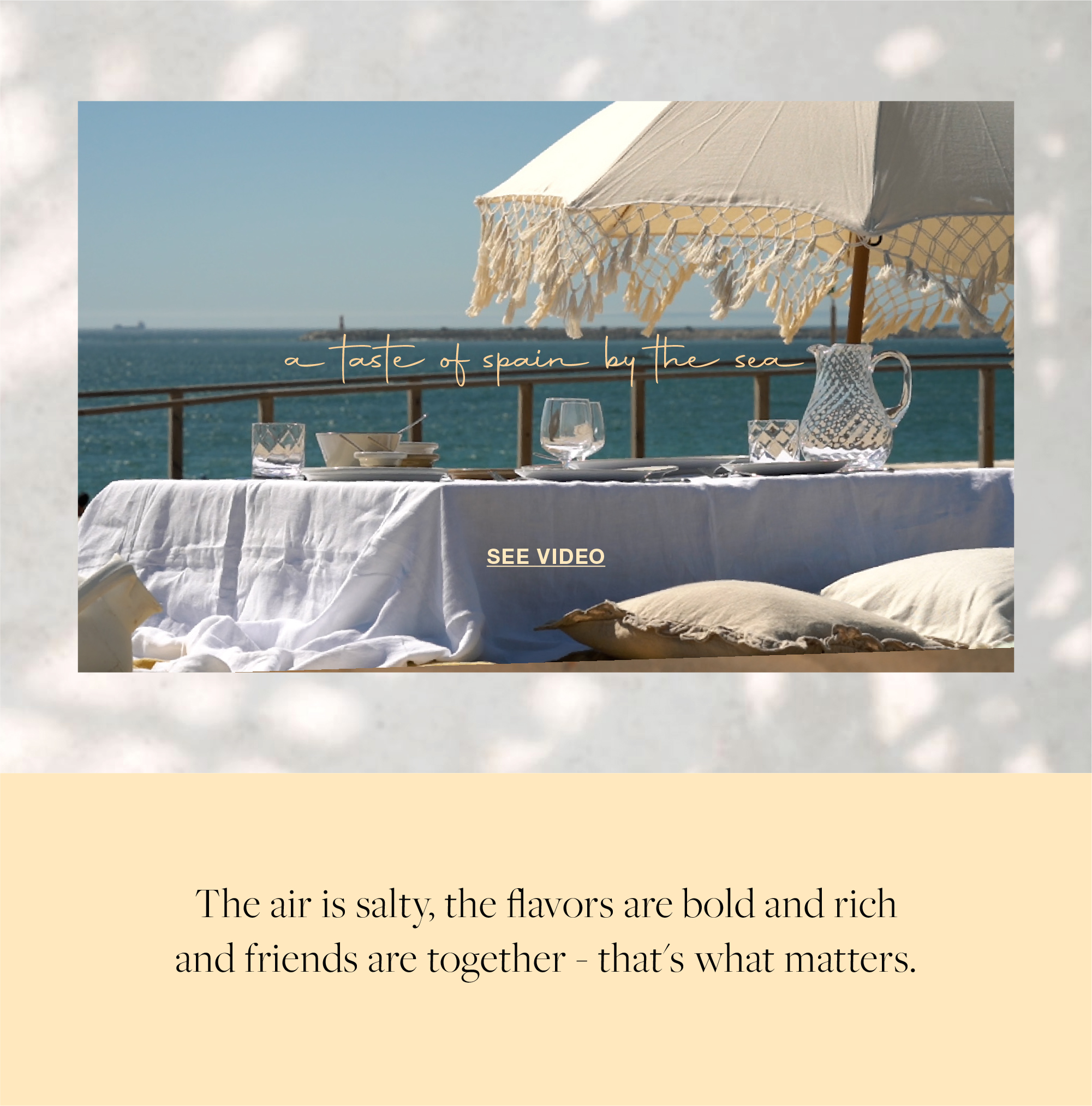 Photograph of a table set by the sea with a white tablecloth, glass pitcher and glasses, a fringed umbrella and the text a taste of spain by the sea and See Video button, followed by text about the salty air, bold and rich flavors and friends together