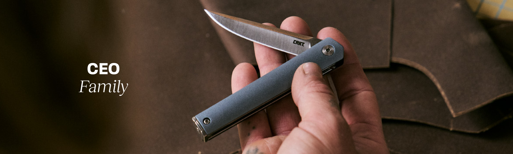 Hand holding an open CRKT CEO folding knife with a slim blue handle against a workshop backdrop