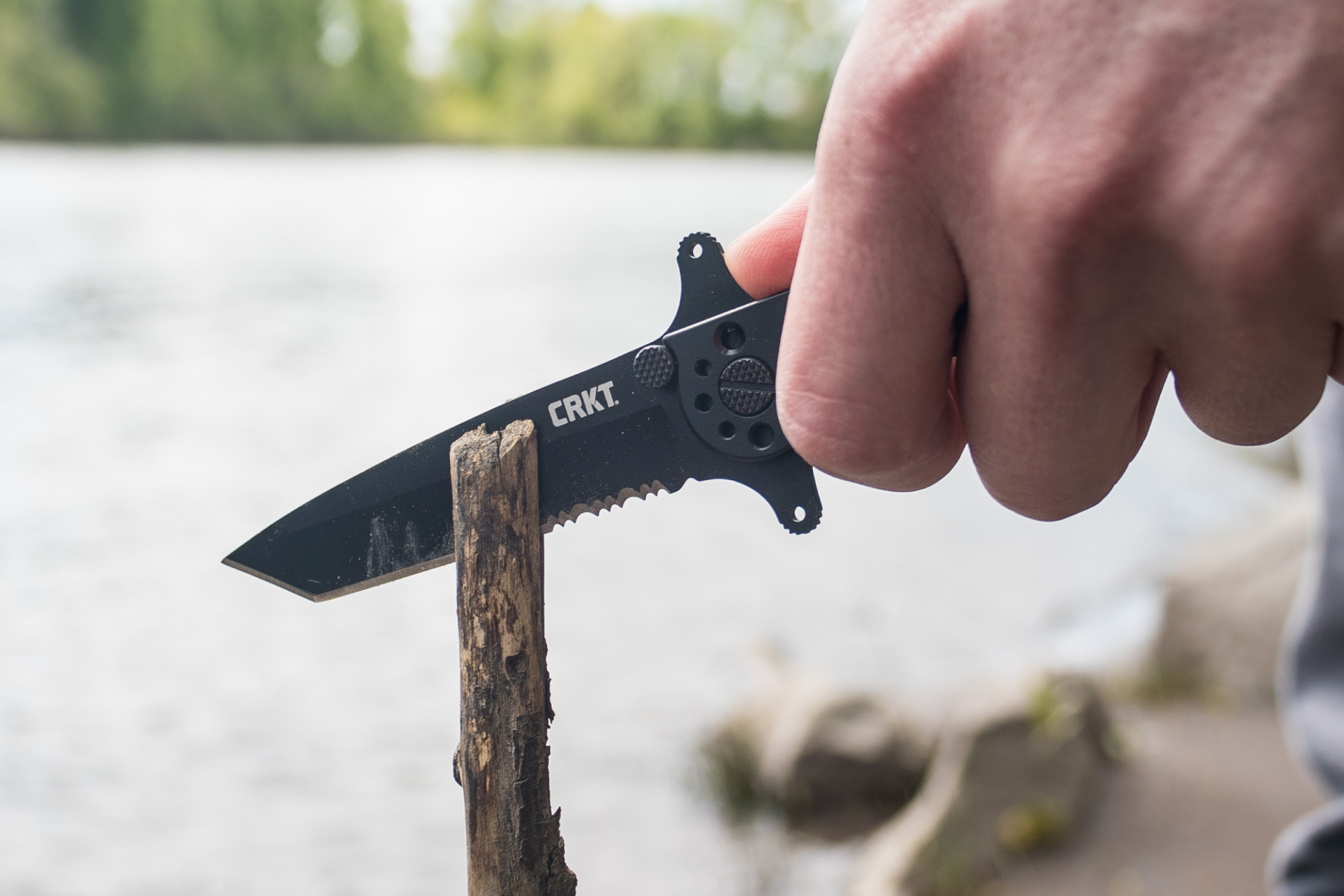 Hand holding a black CRKT M16 Tanto folding knife while cutting into a wooden branch outdoors near a body of water