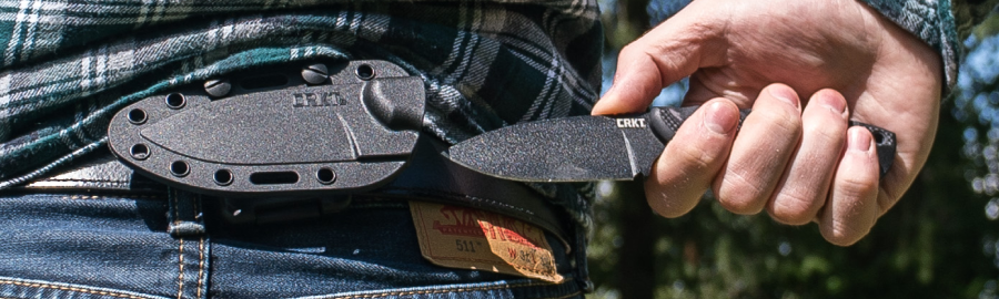 Person drawing a fixed blade CRKT knife from a belt-mounted sheath outdoors.