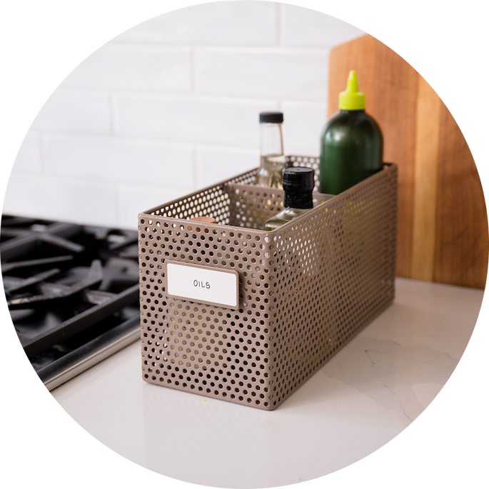 Small perforated basket holding oils and vinegars by the stovetop.