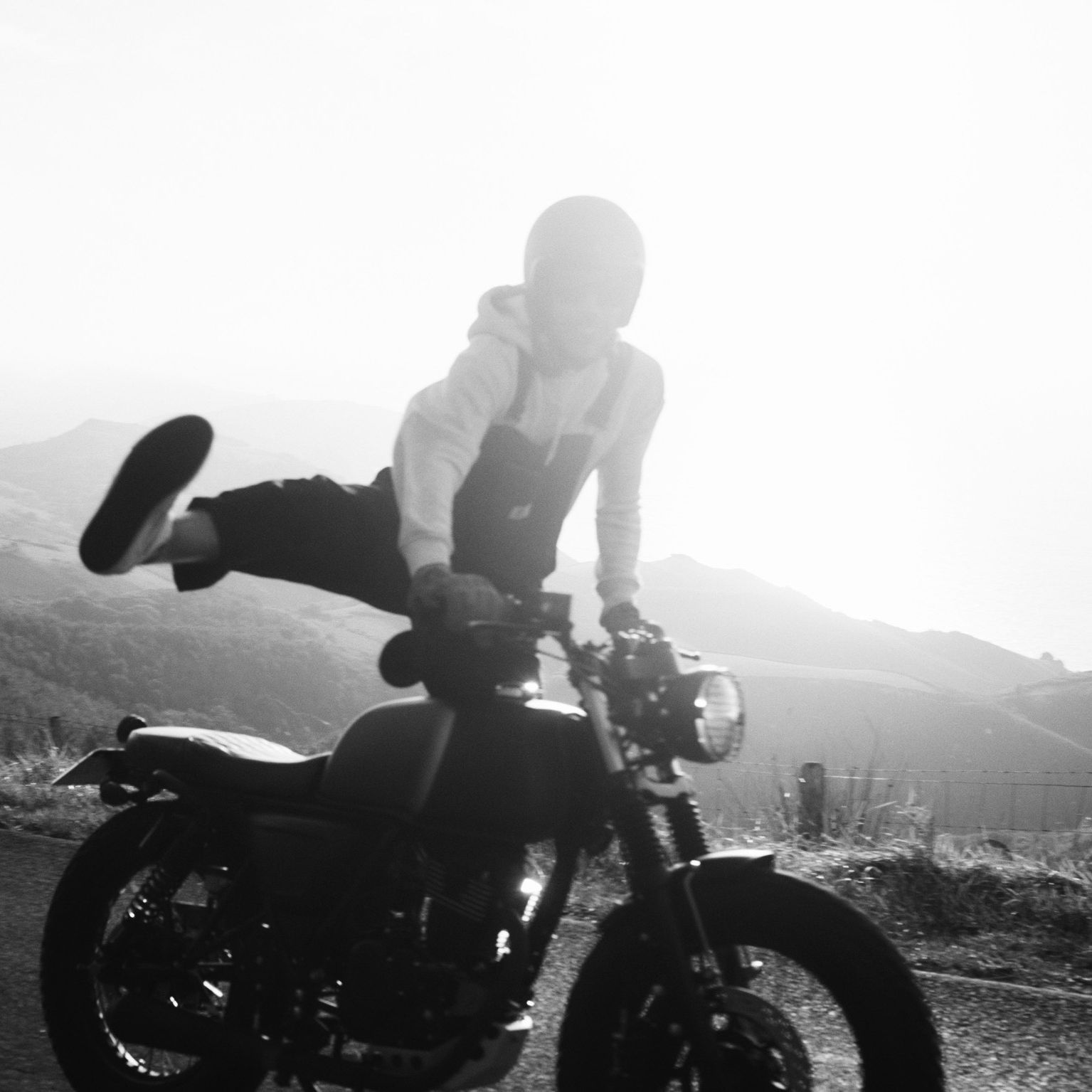 Black and white image of a rider on a Mutt Akita, they're standing on the foot pegs, with one leg up in the air.