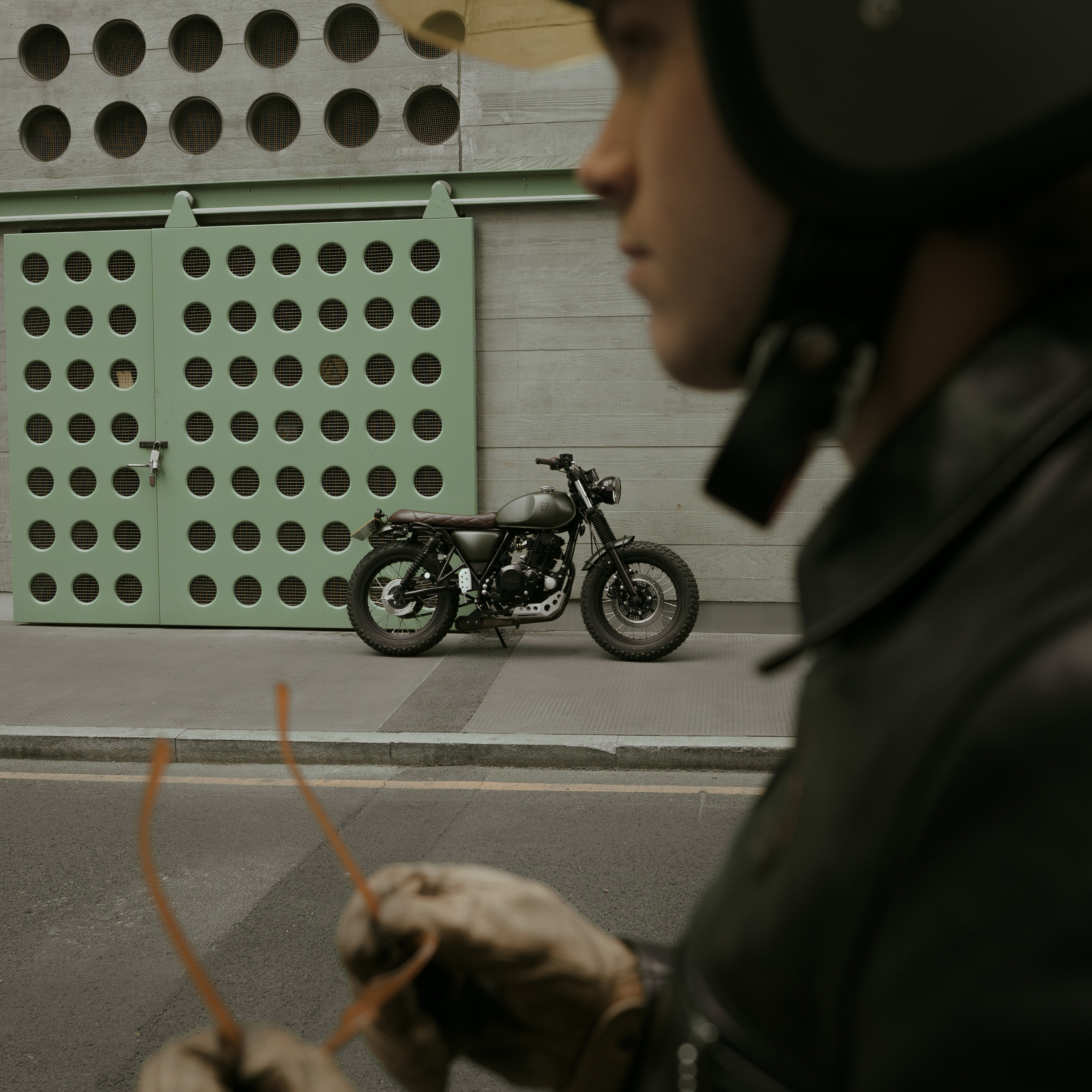 Our Matt Green Mastiff in the centre of the shot, in London, with a light green mesh wall behind it. In the foreground, the rider, wearing a helmet and leather jacket, takes off his orange framed sunglasses.