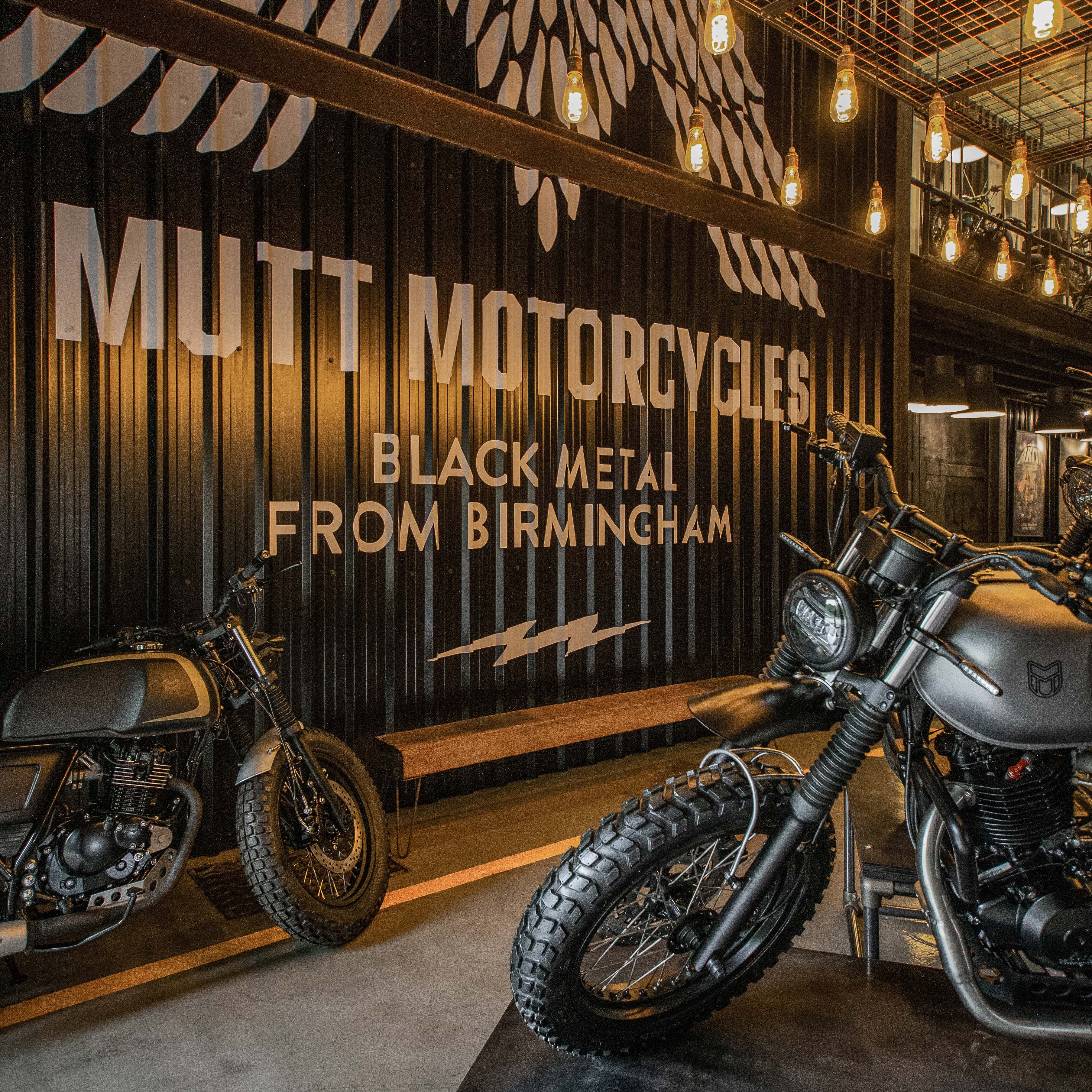 A photo of our Mutt HQ in Birmingham, focusing on the black corrugated steel wall, with "Mutt Motorcycles - Black Metal From Birmingham" in white text, with a large winged M graphic above it. Also in the shot is our Titanium Grey Mushman bike and our Black and Gold Akita bike.