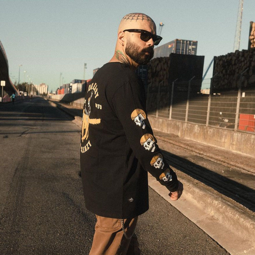 A model wears a Mutt black long sleeve tee the model is wearing, saying "Ride easy, stay sleazy" along with a graphic of a skeleton riding a huge dog. He is also wearing black sunglasses and has a bald head, with a spiders web tattooed on it