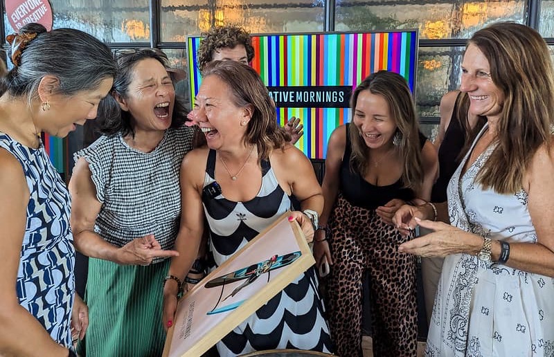 Photo of six women laughing together at a CreativeMornings event, gathered around a colorful striped 'CreativeMornings' backdrop.