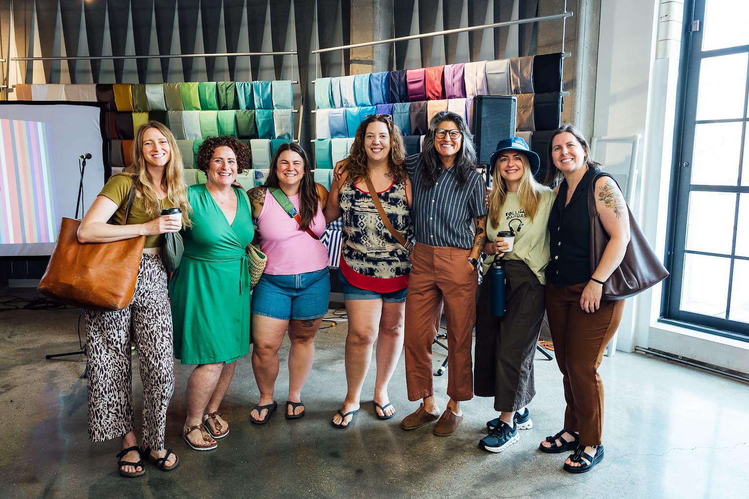 Photo of seven women stand arm-in-arm smiling at the camera in a bright industrial studio space, with a wall of colorful fabric swatches on hanging rods behind them and a striped test print visible on a screen to the left.