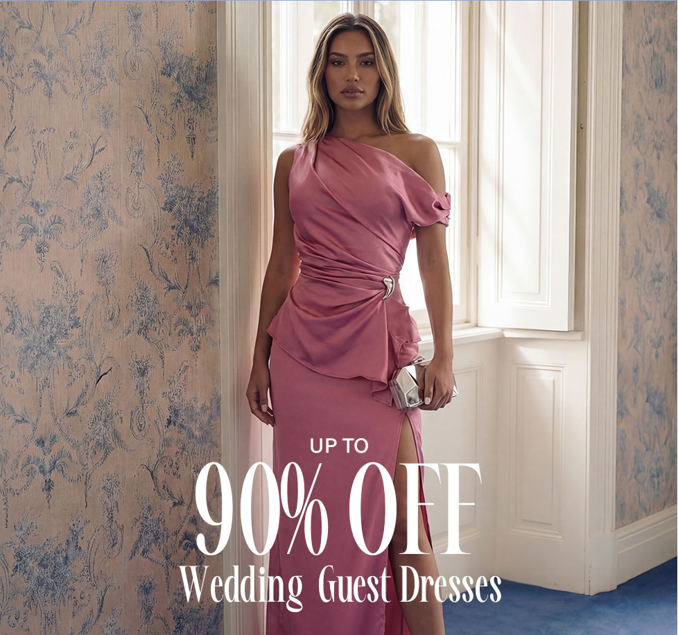 Elegant woman in a flowing pink satin one-shoulder wedding guest dress with a slit, holding a clutch, against a patterned wall, for 90% off sale.
