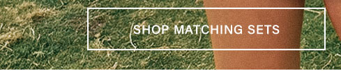 Lower body view of someone in a rustic orange matching set, standing on lush green grass. White button reads: 'SHOP MATCHING SETS' to explore coordinated apparel.