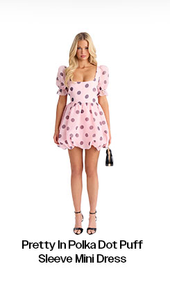 Blonde model showcasing the Pretty In Polka Dot Puff Sleeve Mini Dress in pink with dark spots, complete with heels and handbag.