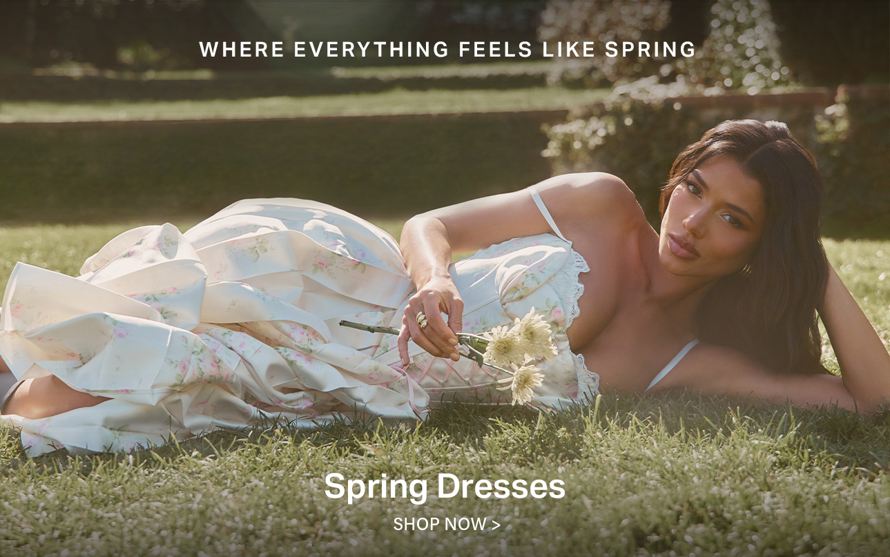 Woman in a flowing cream floral spring dress with ruffled details, lounging on green grass holding white flowers. Discover our new spring collection.