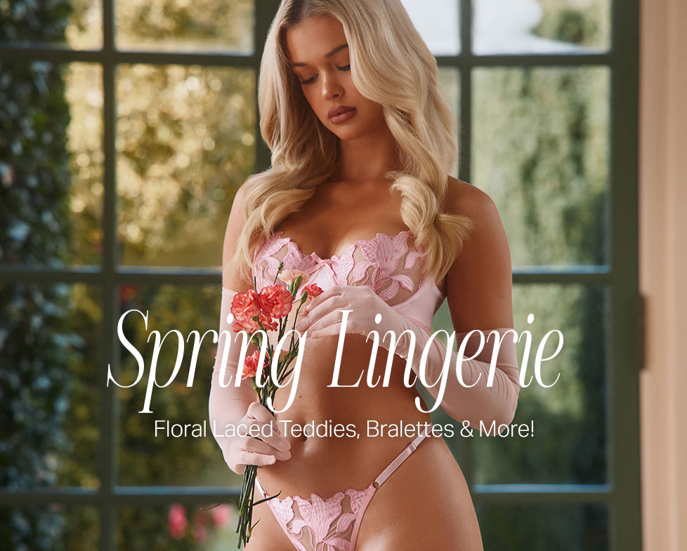 Blonde woman modeling pink floral lace lingerie and long gloves, holding flowers by a window. Showcases spring collection with teddies, bralettes & more.