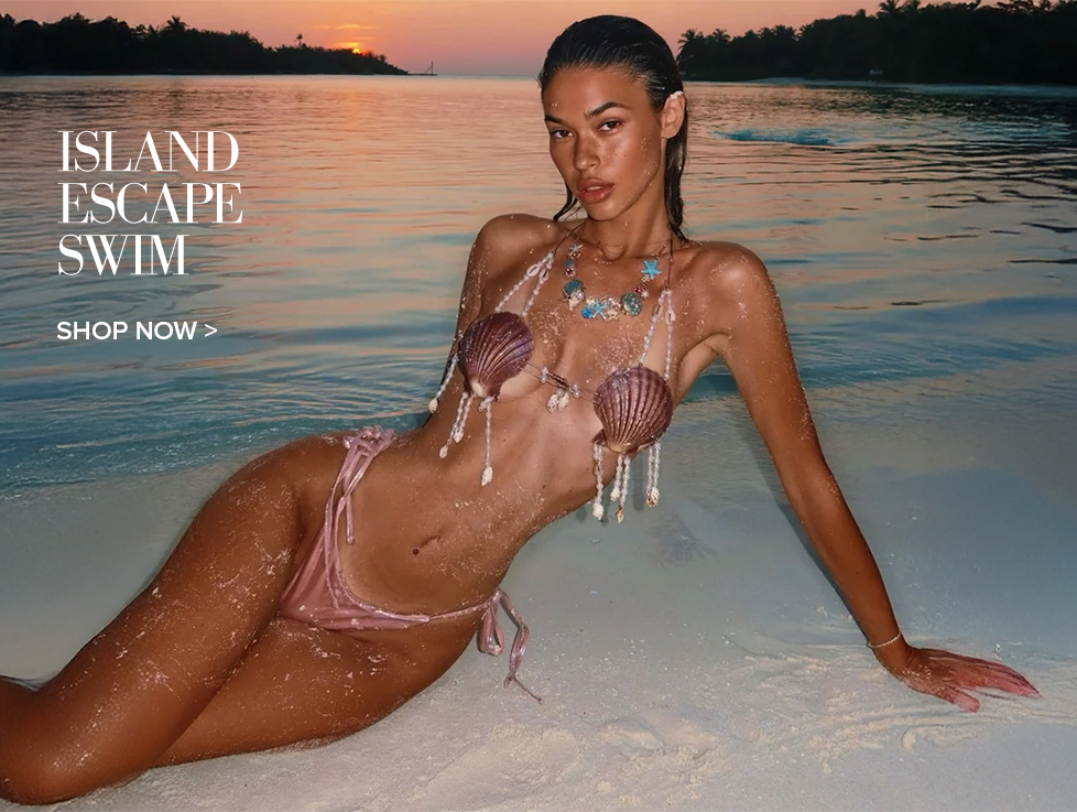 Woman in a unique seashell and beaded top with pink bikini bottoms, reclining in shallow sunset water. Shop Island Escape Swim collection.