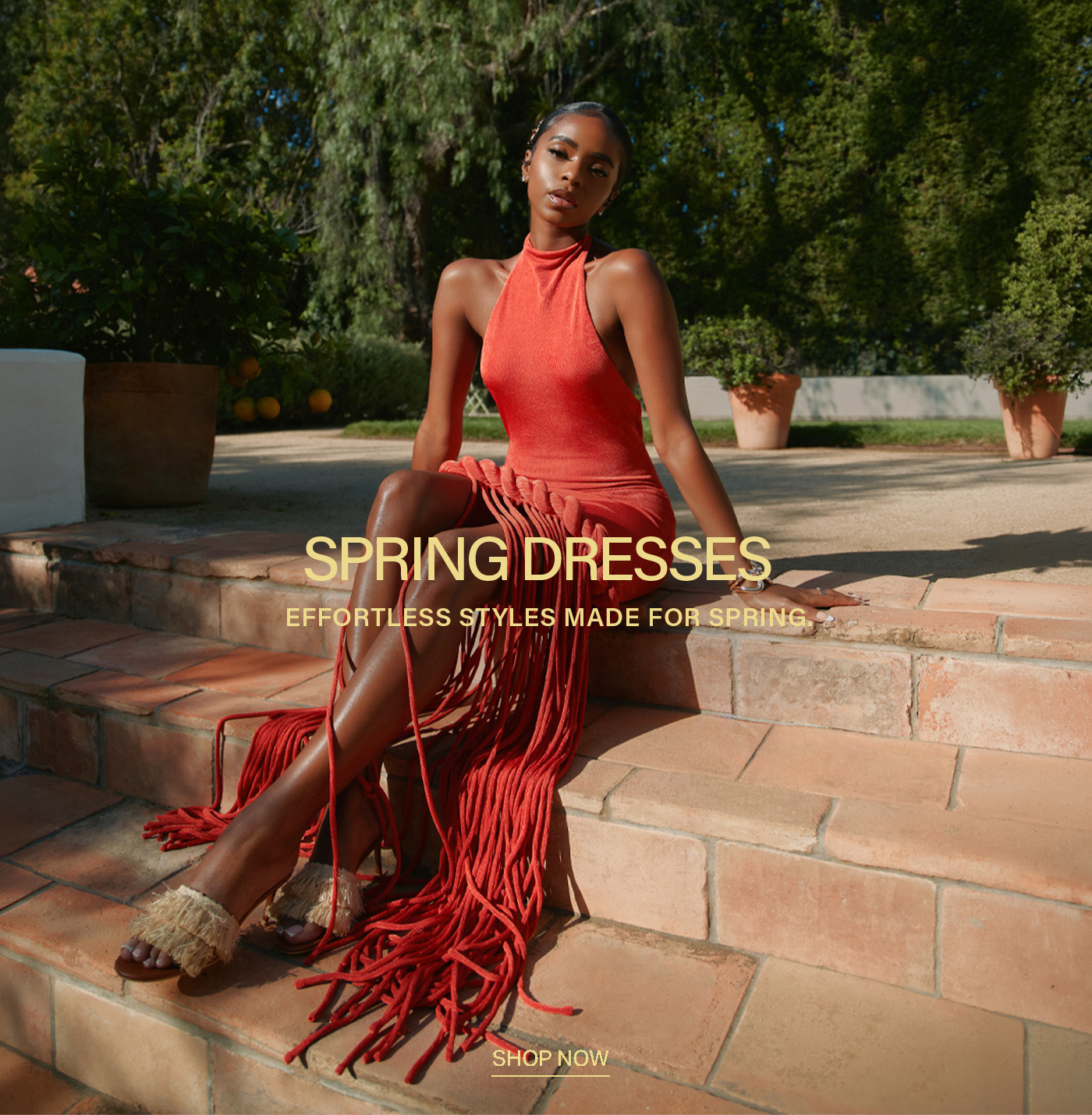 Woman in a vibrant coral halter-neck fringe dress and fringed sandals, sitting on terracotta steps. Discover effortless spring dresses.