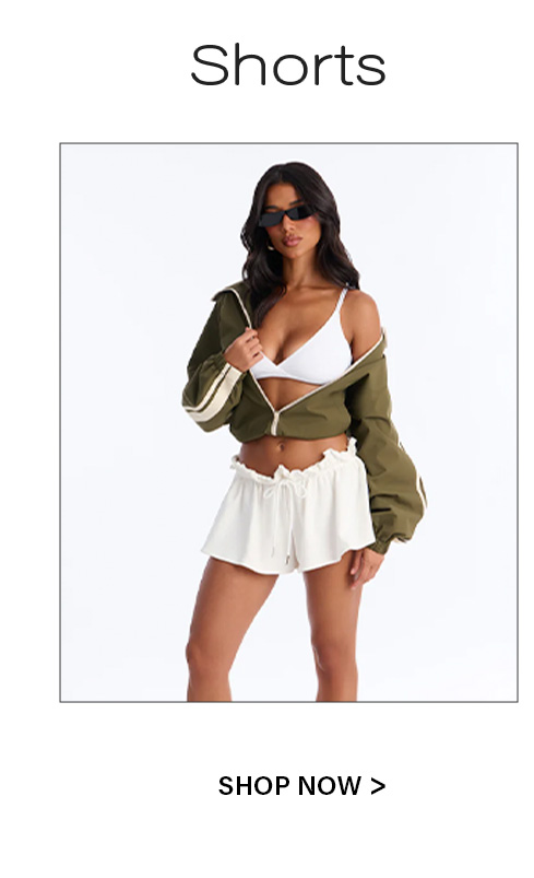 Stylish woman in black sunglasses, white bralette, unzipped olive jacket, and flowy white drawstring shorts, promoting women's shorts.