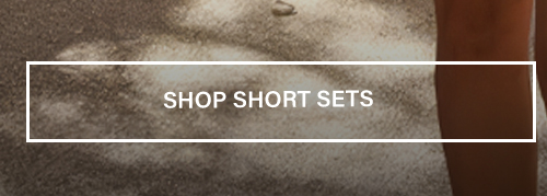 White "SHOP SHORT SETS" button over sunlit sandy ground with a leg, directing users to shop the brand's matching summer short sets.