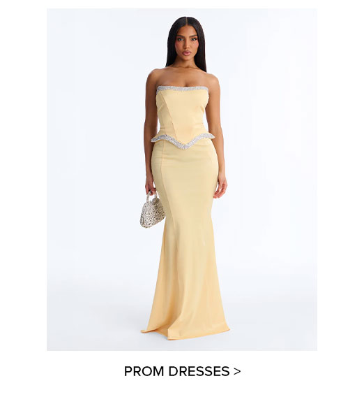 Woman wearing an elegant champagne strapless mermaid prom dress with crystal trim and holding a silver beaded clutch. Explore new formal gowns.