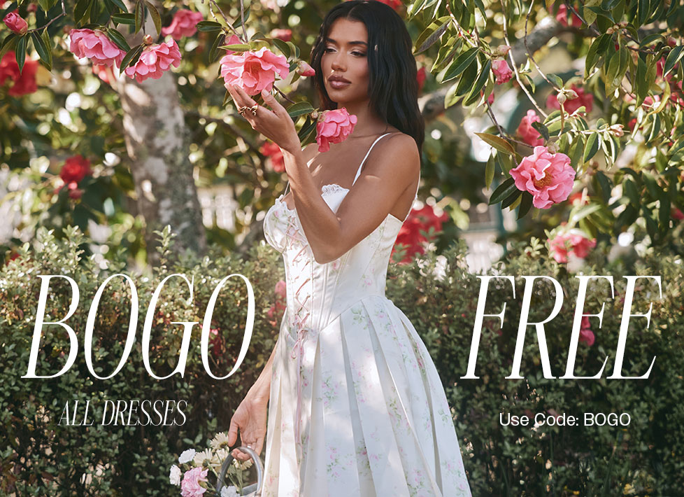 Woman wearing a white floral print spring dress, surrounded by pink garden flowers. BOGO FREE on all dresses - use code BOGO.