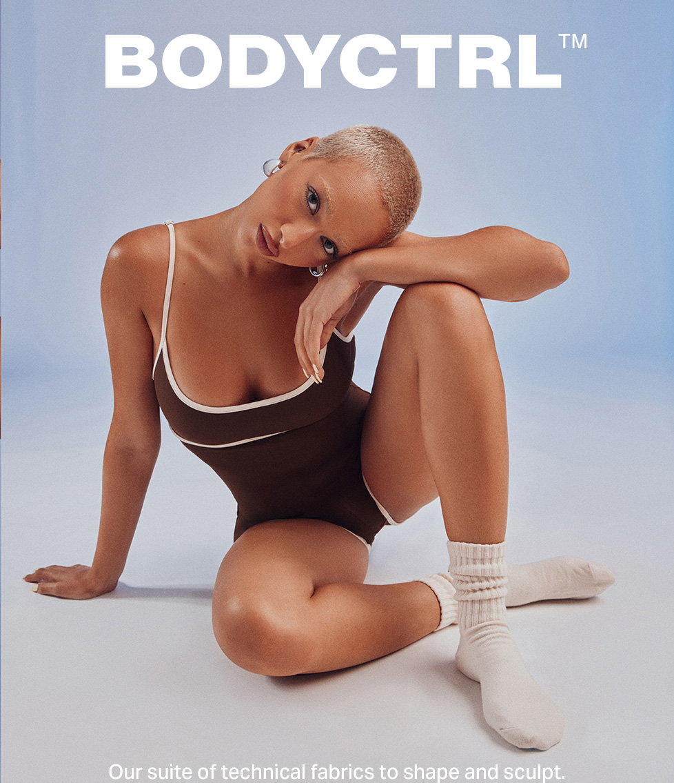Model posing in a brown Bodyctrl sculpting bodysuit with white trim and knit socks, showcasing the brand's new shaping shapewear collection.