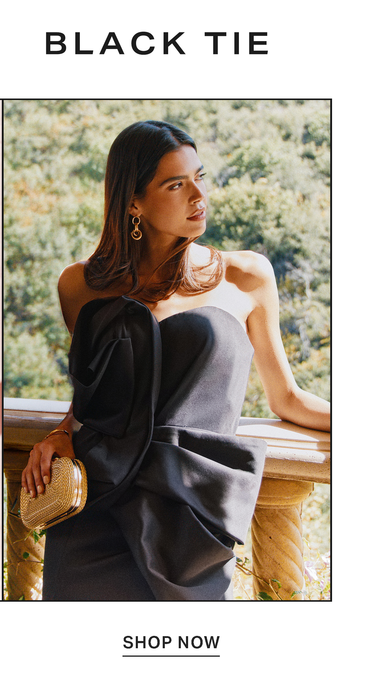 Woman in a strapless black tie dress with a gold clutch on a balcony. Shop now to browse our elegant evening gowns and formal wear.