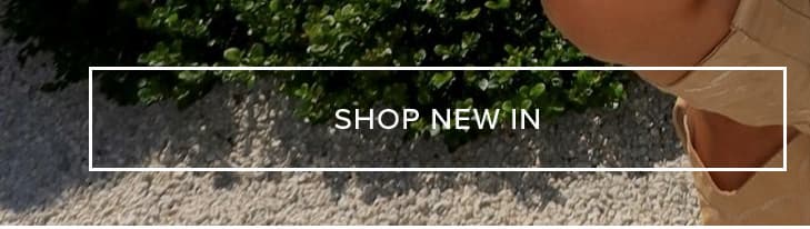 Partial view of a person wearing light beige clothing, set against vibrant green bushes and light gravel. The call to action: "SHOP NEW IN" promotes new collection arrivals.