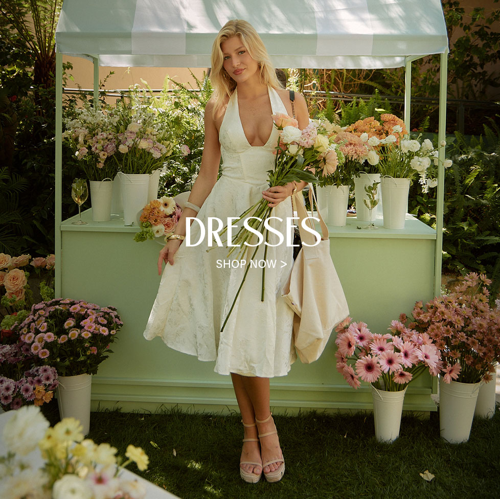 Woman in a white halter dress holding flowers at a mint green floral stand. Promotes elegant summer dresses for women. Shop now.