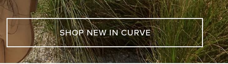 Person modeling a stylish light brown garment, standing gracefully in tall green natural grass. Shop new in curve fashion now.