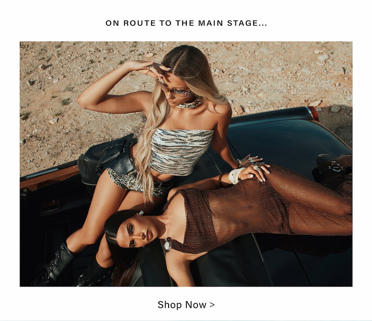 Two women display festival fashion on a black vintage car in the desert. Zebra top, leopard shorts, sheer brown dress. Shop trendy main stage outfits.
