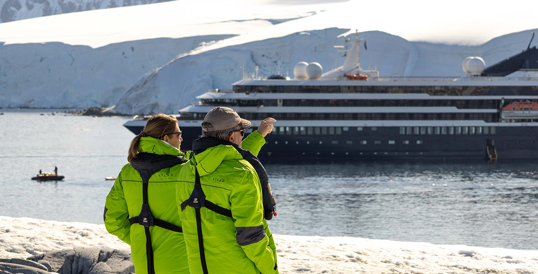 A couple of Atlas Explorers in their Atlas parkas in Antarctica looking out at the yacht at sea from shore