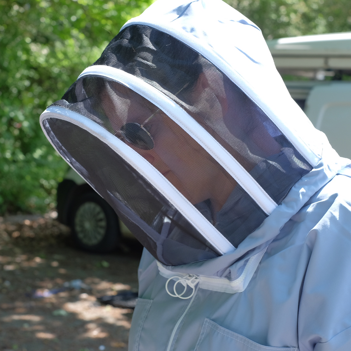 A woman in a blue beekeeping suit with the hood up. The suit is essentially an all in one boiler suit with lots of pockets, and a mesh hood that protects the face