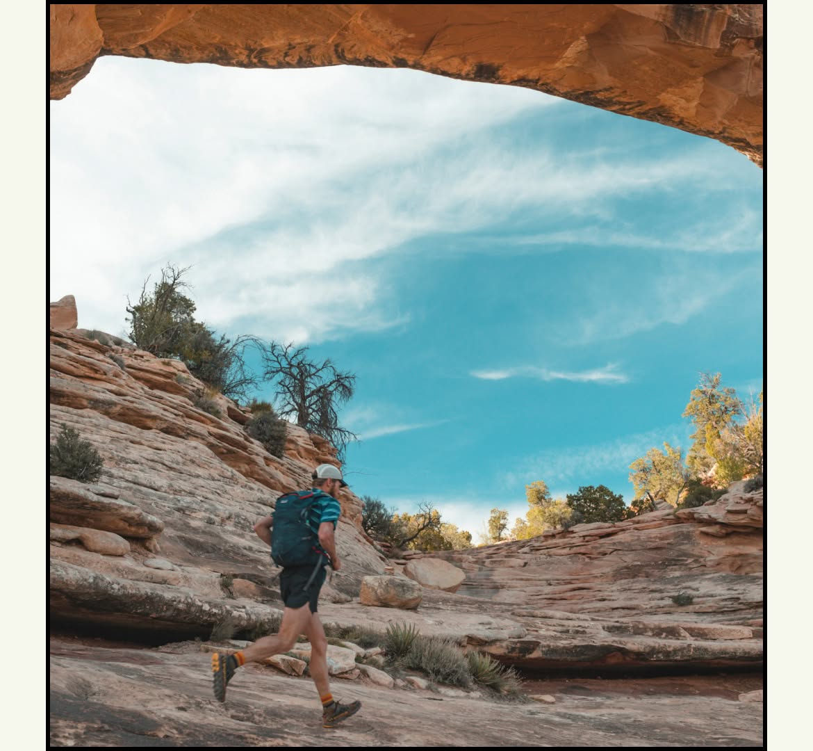 A person wearing a backpack runs on desert lands. A natural sandstone arch frames the sky.
