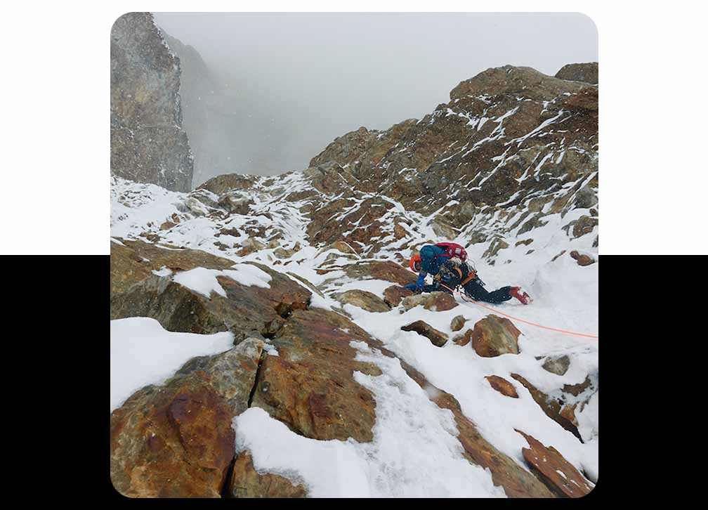 An alpine climber ascends a rocky mountain face covered in snow.